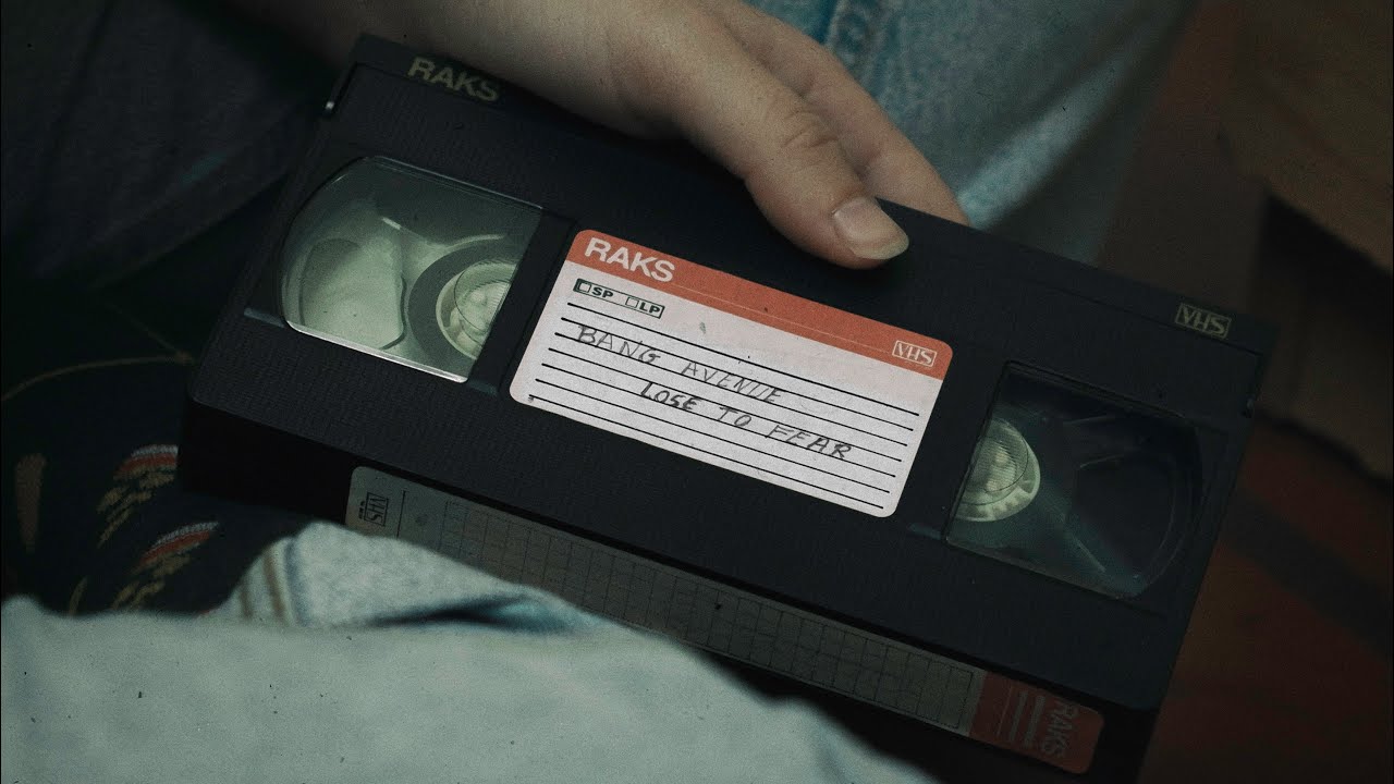 Uma cassete de VHS está pousada nas pernas de uma pessoa. Uma mão agarra a cassete, e nesta última está escrito: "Bang Avenue", "Lose to Fear".